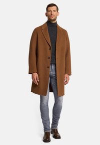 Cappotto di lana marrone con colletto a punta, tre bottoni e tasche laterali, abbinato a un dolcevita scuro, jeans chiari e stivali di pelle marrone.