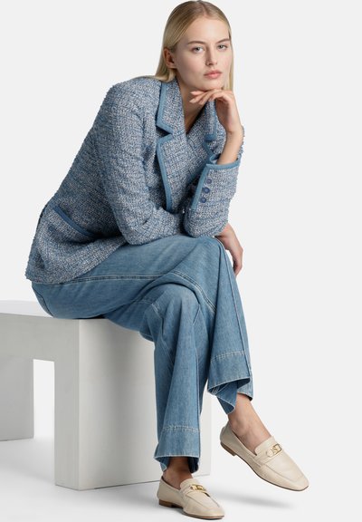 Blazer en tweed bleu clair avec bordure contrastante, associé à un pantalon en denim large bleu clair et des mocassins beiges avec un accent doré.