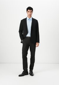Černý blazer přes světle modrou košili, doplněný tmavě šedými kalhotami a černými společenskými botami. Hladká tkanina a dobře padnoucí střih bez viditelných vzorů.