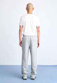 T-shirt en coton blanc et jogging gris clair avec une poche arrière, présentés de derrière sur un tapis bleu contre un fond uni.