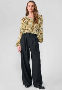 Bedruckte gelbe und grüne Bluse mit lockerer Passform und gerüschtem Ausschnitt, kombiniert mit hoch taillierten schwarzen weit geschnittenen Hosen, glatte Textur.