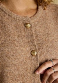 Brązowy dziergany kardigan o subtelnej teksturze, z trzema złotymi guzikami z wytłaczanym wzorem, noszony na dłoni ozdobionej srebrnymi pierścionkami.