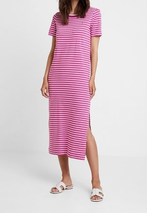 Kobieta ubrana w różowo-białą midi sukienkę w poziome paski z krótkimi rękawami i bocznym rozcięciem, w zestawieniu z białymi sandałami na płaskiej podeszwie.