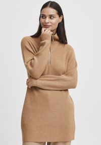 Pull en maille côtelée marron clair avec un col haut et une fermeture éclair semi-longue, arborant un motif en chevrons et des manches longues.