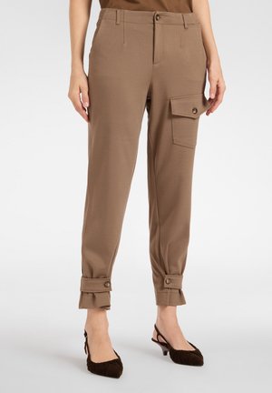 Braune, zulaufende Hose mit geknöpften Knöchelriemen und einer geknöpften Klappentasche am rechten Oberschenkel, getragen mit braunen Slingback-Pumps mit Absatz.