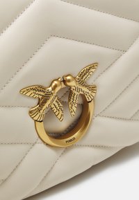 Borsa in pelle beige trapuntata con hardware dorato rotondo che presenta due uccelli che si baciano. Texture liscia e dettagli di cucitura ben visibili.