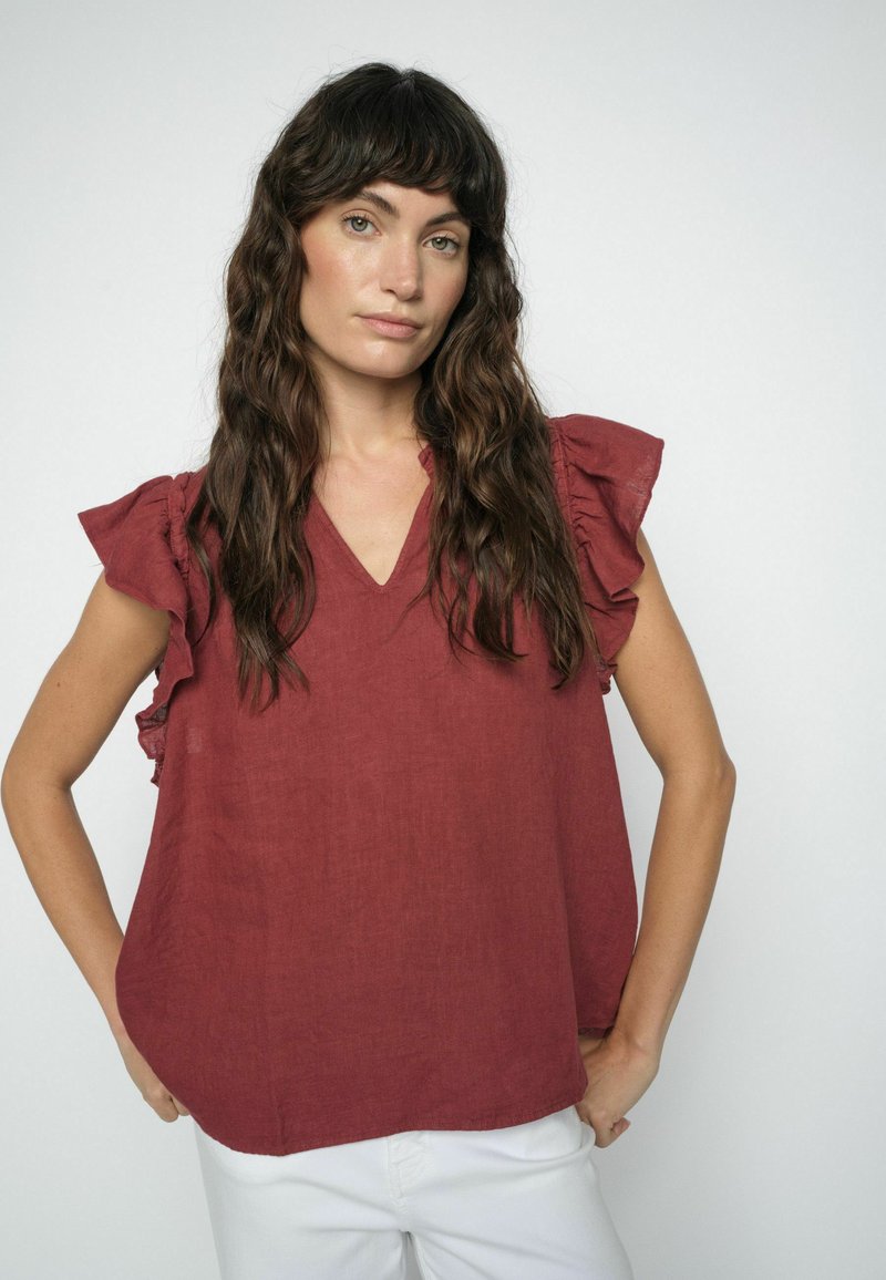 Mujer con cabello largo y ondulado de color castaño, vestida con una blusa sin mangas de color óxido y pantalones blancos, de pie frente a un fondo claro y liso.