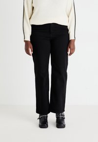 Schwarze weit geschnittene Jeans kombiniert mit einem cremefarbenen Pullover aus strukturiertem Stoff und verzierten Ärmeln; schwarze Schuhe mit Nietenakzenten.