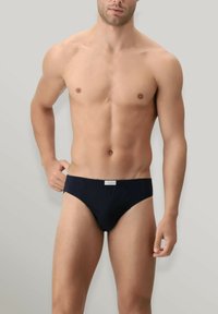 Culotte bikini da uomo nere realizzate in tessuto morbido, con un design semplice, taglio basso e una piccola etichetta con logo in vita.