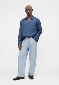 Homme aux cheveux blonds courts portant une chemise bleue ample à manches longues, un jean large bleu clair, une ceinture marron et des chaussures noires, debout avec une main dans la poche.