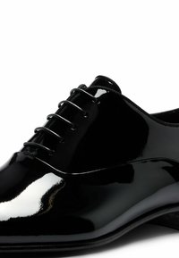 Chaussure habillée en cuir verni noir avec une surface lisse et brillante, un bout arrondi et cinq paires d'œillets pour les lacets.