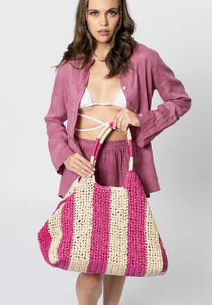 Kobieta w różowej koszuli i szortach trzyma dużą plecioną różowo-kremową torbę w paski z okrągłymi rączkami na jednolitym tle.