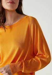 Gros plan d'une personne portant un pull en tricot à manches longues de couleur orange vif, avec un col rond et des poignets côtelés.