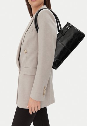 Žena v béžovém blejzru nese přes rameno lesklou černou koženou tašku, zobrazena z profilu na neutrálním pozadí.