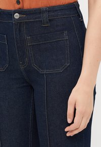 Nahaufnahme einer Hand einer Person, die neben einer dunkelbärtigen Jeans mit sichtbaren Nähten und Knopfdetails am Hosenbund liegt.