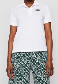 Valkoinen kauluspaita, jossa on kaulus ja vihreä sekä sininen brodeerattu logo. Yhdistetty vihreisiin housuihin, joissa on toistuva valkoinen tekstikuvio.