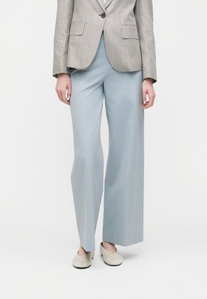 Kobieta ubrana w jasnoniebieskie spodnie o szerokich nogawkach, beżową marynarkę w kratę oraz beżowe buty wsuwane, stojąca na tle jednolitego tła.