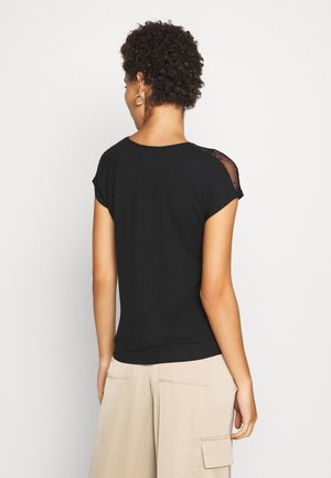 Maglietta nera a maniche corte con scollo rotondo e dettagli in pizzo sulle spalle, indossata con pantaloni cargo beige, vista da dietro.