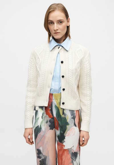 Cardigan crème en maille torsadée avec boutons noirs, superposé sur une chemise à col bleu clair, associé à un pantalon à motif floral coloré.