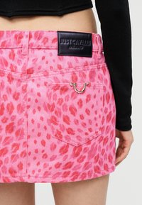 Κοντινό πλάνο ενός ατόμου που φοράει ροζ denim φούστα με μοτίβο λεοπάρδαλης, με μαύρη ετικέτα Just Cavalli Milano και ασημένιο έμβλημα σε σχήμα πέταλου στην πίσω τσέπη.