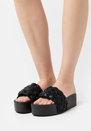 Mustat platform-sandaalit, joissa on letitetty nahkaosa, avoin varvashuone ja teksturoidun pohjan. Pohjassa on pieni viistemuoto nostetta varten.
