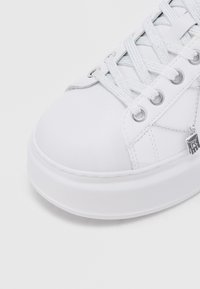 Λευκό αθλητικό παπούτσι με λείο δερμάτινο επάνω μέρος, στρογγυλή μύτη και παχύ λευκό καουτσούκ σόλα. Διαθέτει ασημένια ρυγχοςύνδετα και αντίστοιχους λαμπερούς κορδόνια.