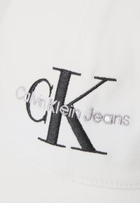 White cotton cap featuring embroidered "Calvin Klein Jeans" logo in black and grey, with a structured shape and curved brim.