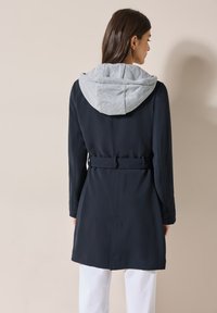 Námořnický modrý trenčkot s šedou mikinou, páskem v pase a dlouhými rukávy. Hladká tkanina se strukturovaným střihem a minimalistickým designem.