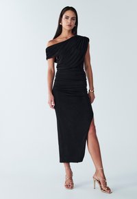 Czarna sukienka na jedno ramię z marszczonym materiałem, bocznym rozcięciem i dopasowanym krojem. Połączona z złotymi sandałkami na paski.