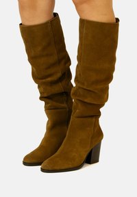 Stivali alti in suede marrone con design morbido, punta quadrata e tacco spesso nero. Dettaglio con zip laterale per una facile calzata.