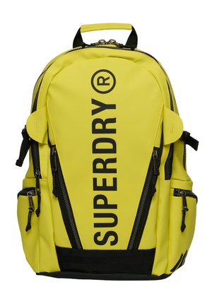 Felgele rugzak met zwarte ritsen en riemen, groot verticaal "SUPERDRY" logo in zwart op het voorste middenpaneel.