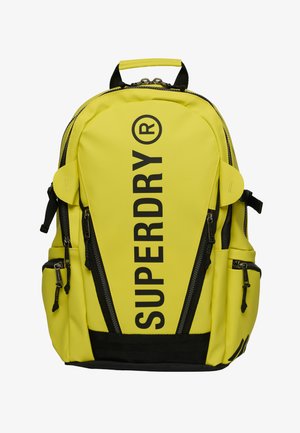Felgele rugzak met zwarte ritsen en riemen, groot verticaal "SUPERDRY" logo in zwart op het voorste middenpaneel.