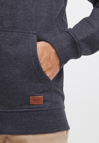 Sudadera gris oscuro con un bolsillo frontal, puños acanalados y una etiqueta de cuero con la inscripción "BLEND" en el dobladillo. Textura suave y diseño ajustado.