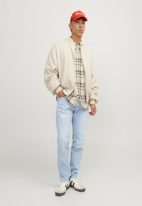Beige bomberjack, geruite shirt, lichtblauwe distressed jeans en witte sneakers met zwarte accenten. Rode pet met logo.