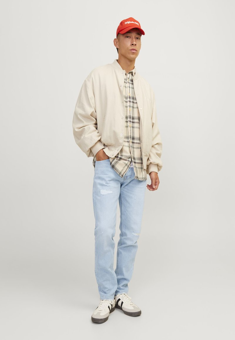 Beige bomberjack, geruite shirt, lichtblauwe distressed jeans en witte sneakers met zwarte accenten. Rode pet met logo.