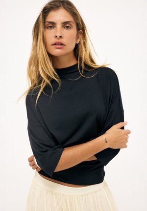 Blondína s rozopnutými vlasmi, nosiaca čierne tričko s vysokým golierom a rukávmi po lakte a svetlú plisovanú sukňu, ruky prekrížené.