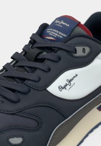 Mørkeblå sneakers med en hvit logo, med mesh- og skinnteksturer, flate lisser og en kremfarget såle med røde og blå detaljer.