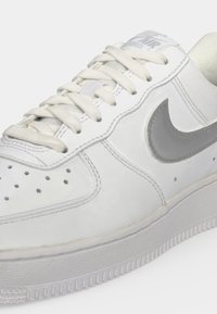 Άσπρο sneaker Nike Air Force 1 με μπεζ κορδόνια, διάτρητη περιοχή δακτύλων και γκρι λογότυπο Nike swoosh στο πλάι.