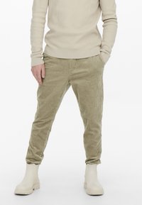 Pantalones de pana oliva claros con un corte ajustado, que presentan canalones verticales y bolsillos laterales, combinados con botas de tobillo de goma beige.