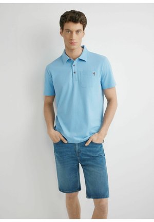 Młody mężczyzna w jasnoniebieskiej koszulce polo z kieszenią i niebieskich dżinsowych szortach, stojący z rękami w kieszeniach na białym tle.