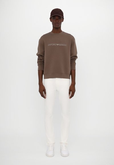 Brown sweatshirt with "Emporio Armani" logo, white fitted pants, and white sneakers. Model also wearing a matching brown cap.