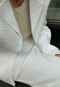 Bílý texturovaný blejzr a odpovídající kalhoty vyrobené z tkaného materiálu, s jemným mřížkovým vzorem a volným střihem. Doplněno béžovým topem.