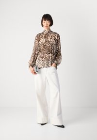 Femme portant un blouse en dentelle à motifs léopard, un pantalon blanc à jambes larges, des chaussures noires à talons pointus, tenant une pochette en argent, se tenant devant un fond uni.