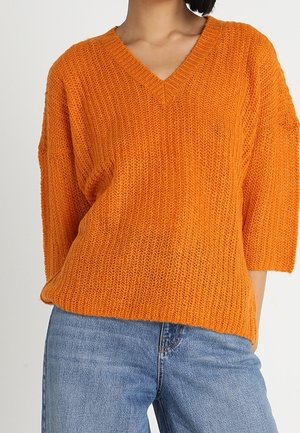 Frau trägt einen strukturierten, orangefarbenen Pullover mit V-Ausschnitt, dreiviertel Ärmeln und blauen Jeans vor einem einfarbigen Hintergrund.
