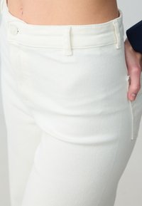 I pantaloni in denim bianco presentano un design aderente con chiusura a bottone e cerniera, passanti per cintura e tasche laterali. Texture liscia visibile.