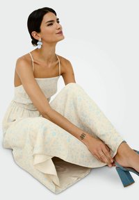 Robe florale jaune clair avec des accents bleus, tissu texturé, bretelles spaghetti et jupe évasée. Associée à des sandales à talons carrés bleues.