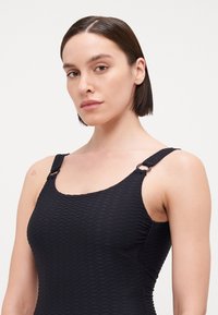 Maillot de bain noir texturé avec décolleté carré, fines bretelles et accents en métal. Le tissu présente un motif en diamant et une finition lisse.