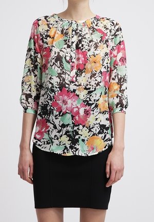 Femme portant un chemisier à manches longues à motifs floraux avec des fleurs roses, orange, jaunes et vertes et une jupe noire au genou.