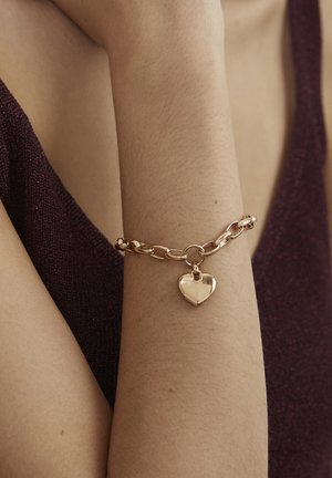 Gold metal bracelet with interlinked chain design and a shiny heart charm, worn on a bare arm against a dark fabric background.