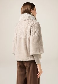 Giacca in pelliccia sintetica color beige chiaro con motivo a treccia testurizzato sulla parte superiore della schiena, maniche larghe e polsini a coste. Pantaloni marroni visibili.